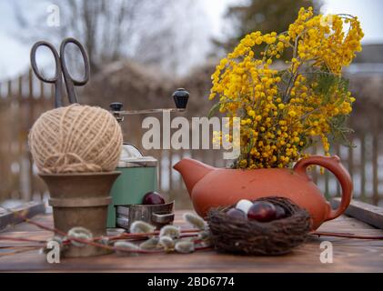 On the wooden table top is a nest with painted quail eggs, willow branches and various household utensils.  Stock Photo