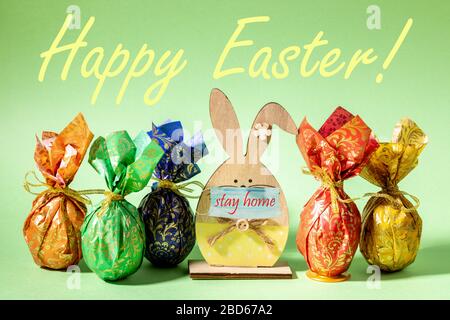 Wrapped in colored paper Easter eggs and wooden figure bunny in mask with words stay home are standing on neo mint backdrop. Words Happy Easter. Pande Stock Photo