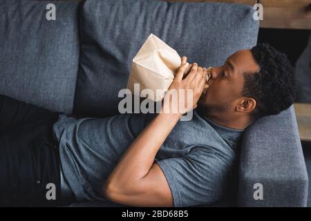 stressed african american man breathing with paper bag while having panic attack at home Stock Photo