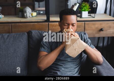 stressed african american man breathing with paper bag while having panic attack at home Stock Photo