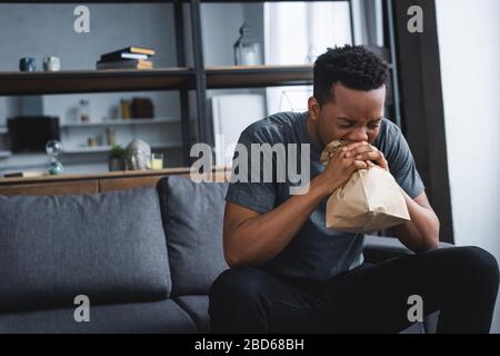 stressed african american man breathing with paper bag while having panic attack at home Stock Photo