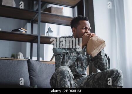 stressed african american soldier in military uniform breathing with paper bag while having panic attack and suffering from PTSD at home Stock Photo