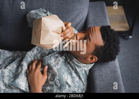 stressed african american soldier in military uniform breathing with paper bag while having panic attack and suffering from PTSD at home Stock Photo