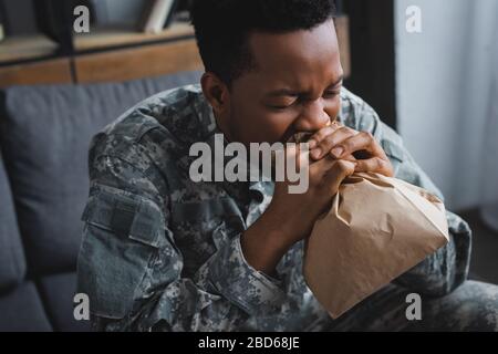 stressed african american soldier in military uniform breathing with paper bag while having panic attack and suffering from PTSD at home Stock Photo