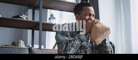 panoramic shot of african american soldier in military uniform breathing with paper bag while having panic attack and suffering from PTSD at home Stock Photo
