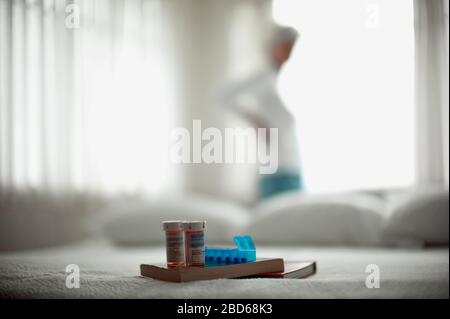 Two pill bottles,  a medication dispenser and two books sitting on a bed with a woman standing in front of a window in the background. Stock Photo