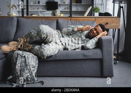 stressed african american soldier in military uniform talking on smartphone and suffering from PTSD at home with backpack Stock Photo