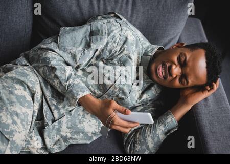 lonely african american soldier crying while holding army badge and smartphone, suffering from PTSD at home Stock Photo
