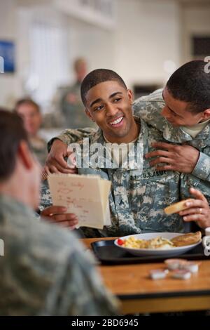 Young soldier looks over his friend's shoulder as he reads a  letter from his young son while eating breakfast in the army mess hall. Stock Photo