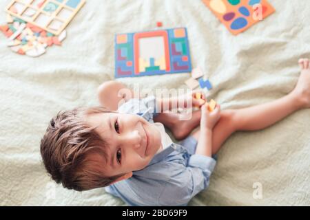 The happy boy sits on the bed and lays puzzles. View from above, child looks at the camera Stock Photo