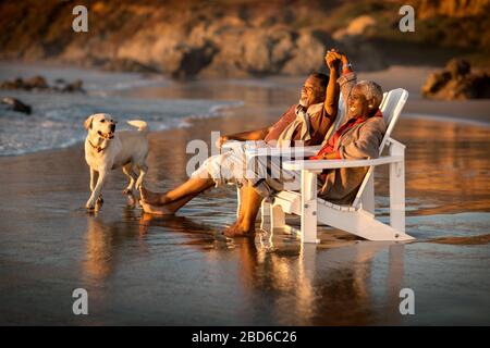 Mature couple relaxing in deck chairs on a beach at sunset. Stock Photo
