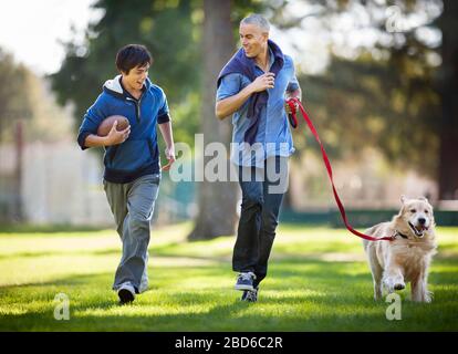 Smiling mid adult man and his son having fun jogging together in the park with their dog. Stock Photo