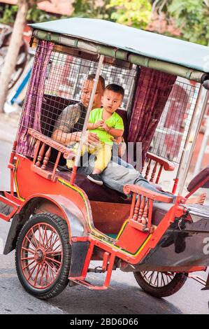 A father holds onto his baby as they sit in the back of a traditional rickshaw and make their way through the busy streets of Phnom Penh. Stock Photo