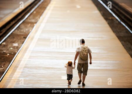 Father holds is toddler's hand as they walk along a platform between train tracks. Stock Photo