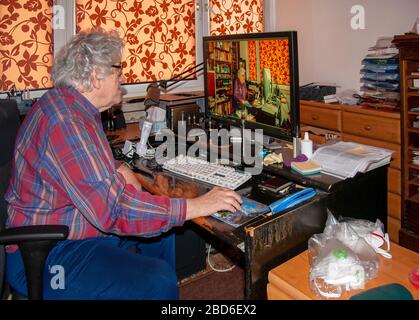 Elderly senior photographer working at home editing a photo with himself working at home on a desktop computer in a mess with antiviral masks ready Stock Photo