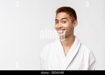 Beauty, spa and leisure concept. Cheeky curious handsome asian man in bathrobe look camera surprised and intrigued, smiling, head interesting proposal Stock Photo
