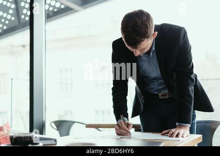 Confident young business man signing documents in a modern office with window in background. Pen in hand, papers on the wooden desk Stock Photo