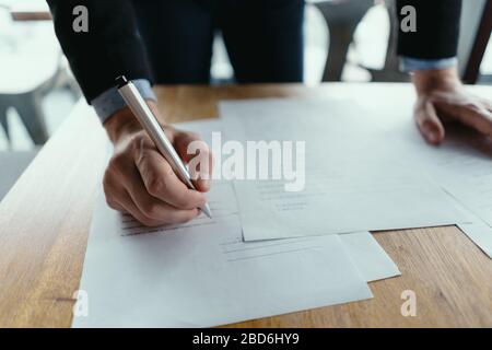 Close up hands signing documents in a modern office with window in background. Pen in hand, papers on the wooden desk, futuristic background. Stock Photo
