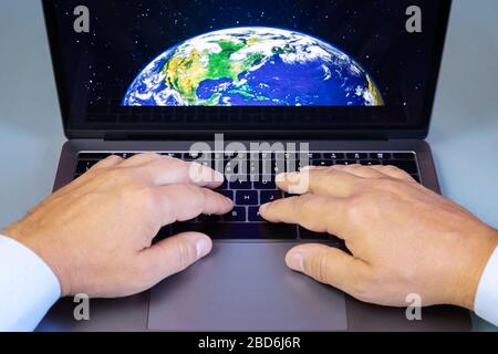 A man types on the keyboard of an Apple laptop, Macbook pro series. The image of the planet Earth seen from space appears Stock Photo