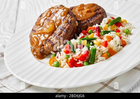 close-up of a portion of grilled ground beef patties with onion gravy and rice mixed with vegetables on a white plate on a wooden table, horizontal vi Stock Photo