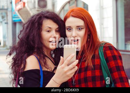 Closeup portrait two surprised girls looking at phone discussing latest gossip news Young shocked funny women friends reading sharing social media new Stock Photo