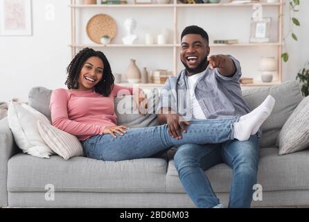 Cheerful black couple watching comedy movie on tv and laughing Stock Photo