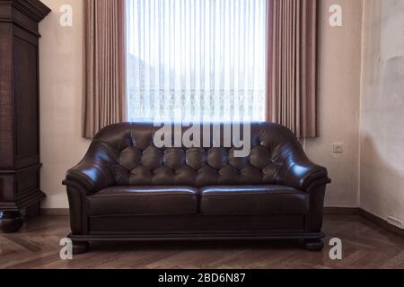 Old leather brown sofa near the window in vintage living room, antique retro design. needs renovation Stock Photo