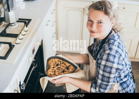 Young blonde woman taking heart shaped berry pie out of the oven. Cooking at home and smiling while looking at the camera. Stock Photo