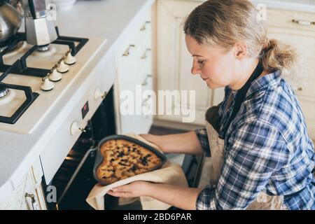 Young blonde woman taking heart shaped berry pie out of the oven. Cooking at home and smiling. Stock Photo