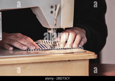 Woman hands using the sewing machine to sew the face medical mask during the coronavirus pandemic. Homemade DIY protective mask against the virus. Sewing masks, COVID-19. Stock Photo