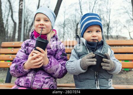 Two cute adorable siblings children sitting on wooden bench and drinking hot chocolate, tea or cocoa from paper cups during walk at city street park Stock Photo