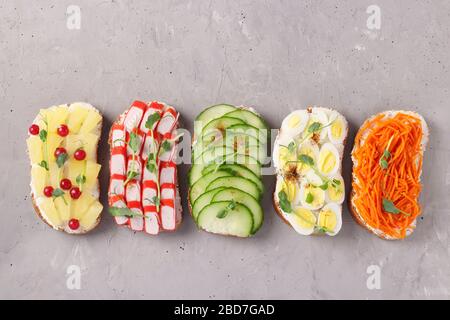 Five sandwiches on toast with fresh carrots, cucumbers, pineapple, red currant, crab sticks and quail eggs with peas microgreens on gray background Stock Photo