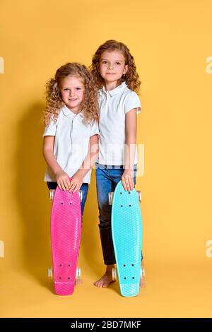 Two Active and happy girls with curly hair having fun with penny board, smiling face stand skateboard. Penny board cute skateboard for girls. Lets ride. Girl with penny board yellow background. Stock Photo