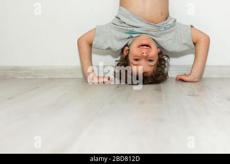 Little smiling cute child having fun standing by hands upside down head feeling positive emotion laughing funny baby child rejoicing on white wall