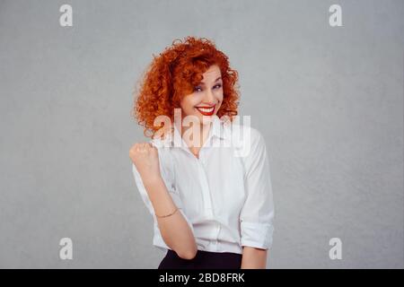 Happy winner. Closeup half body portrait redhead curly woman in white formal shirt exults pumping fists ecstatic celebrates success isolated gray back Stock Photo