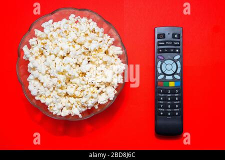 Bowl with salted popcorn next to a tv remote on a red background. Popcorn and tv control concept. Stock Photo