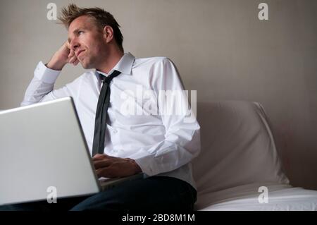 Bored man in smart casual jeans sitting on the sofa browsing his laptop computer Stock Photo
