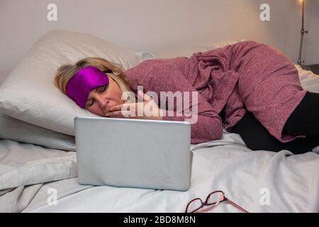 woman in bed with bathrobe and eye mask reading on her computer and yawning Stock Photo