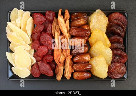 Сandied slices of ginger root and strawberries, dried pear slices, dates, peaches and apricots lying on a black ceramic plate on a black stone surface Stock Photo