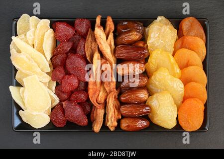 Сandied slices of ginger root and strawberries, dried pear slices, dates, peaches and apricots lying on a black ceramic plate on a black stone surface Stock Photo