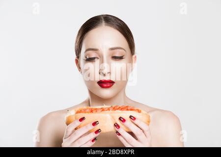 Not healthy. Young beautiful woman wants to eat hotdog craving but she stops because it is unhealthy food. Natural red lips black eyeliner girl, false Stock Photo