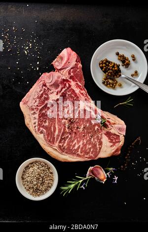 Bone-in raw marbled pork steak with spice rub, fresh herbs and garlic for seasoning in a flat lay on a dark background Stock Photo
