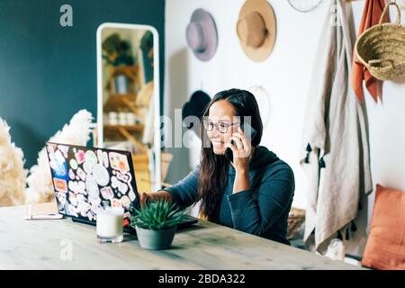 Young charming stylish woman in a home office sits at a desk, looks at a laptop and speaks while smiling on the phone. Stock Photo
