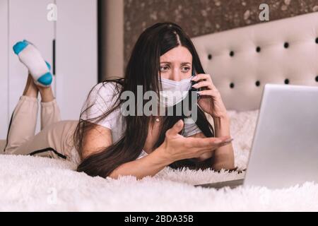 Girl in a mask works on a laptop at home in isolation. masked girl is working on a laptop at home in quarantine. Work on remote access. Self isolation Stock Photo