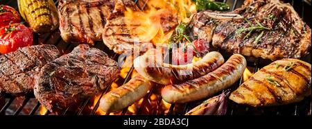 Close-up banner with various delicious kinds of pork meat and sausages on barbecue burning grill with fresh tomatoes, corn and spicy herbs Stock Photo