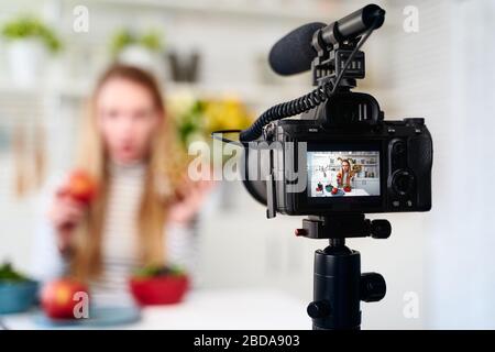 Display of camera recording video blog for food blogger woman with apple, pineapple in kitchen studio talking about healthy vegan eating. Influencer Stock Photo