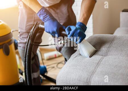 Cleaning service. Man janitor in gloves and uniform vacuum clean sofa with professional equipment. Stock Photo