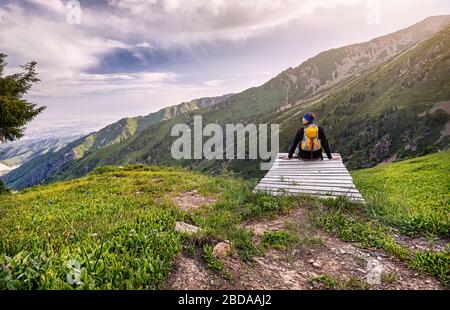 Hiker in with yellow backpack sitting on the bench with view of mountains at sunrise sky background Stock Photo