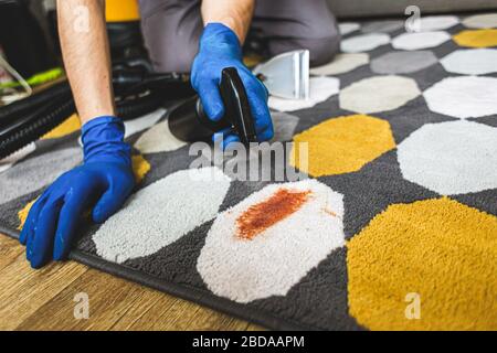 Close-up Of Person's Hand Cleaning Stain On Carpet With Sponge. Cleaning service. Man janitor in gloves and uniform vacuum clean carpet with professio Stock Photo