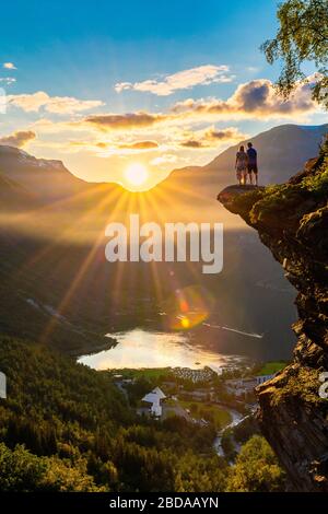 Man and woman admiring sunset over Geiranger village and Geirangerfjord from top of rocks, More og Romsdal county, Norway Stock Photo